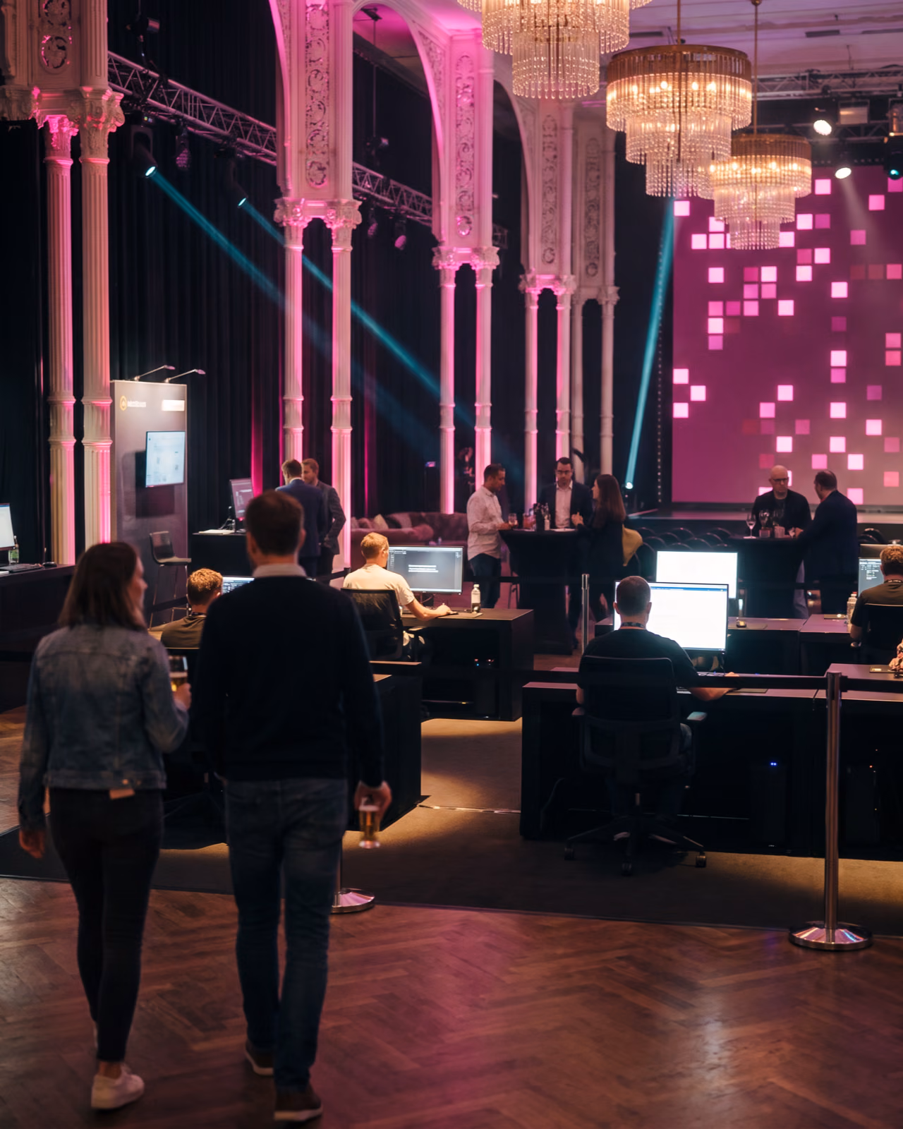Gäste im Festsaal der Flora bei Nacht, dicht an den Bildschirmen der Galerie, dahinter die Bühne im Magenta-Licht. KI-Visualisierung des geplanten Abends. Basis: echtes Foto der Flora Köln.