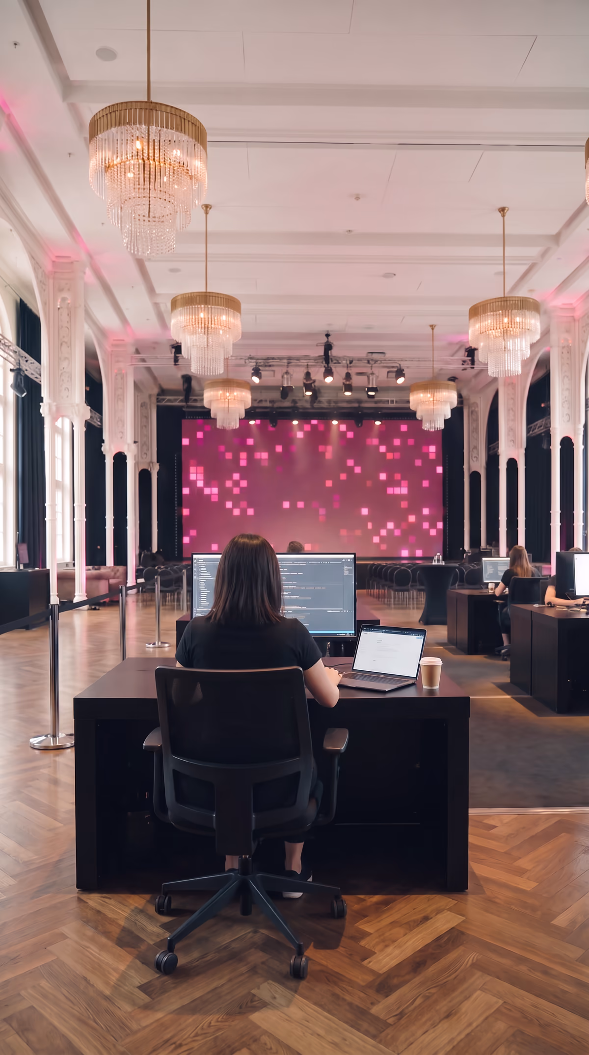 Über die Schulter eines Solo-Startplatzes in der Flora: Monitor, Laptop und Code, dahinter ein Teamtisch, im Hintergrund die Bühne bei Tageslicht. KI-Visualisierung des geplanten Tages. Basis: echtes Foto der Flora Köln.